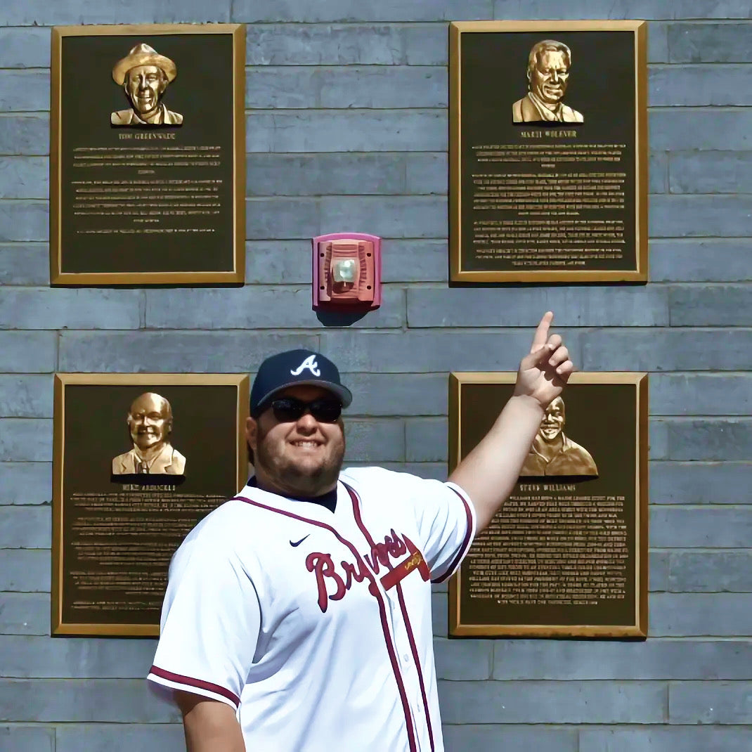 Man in a baseball jersey pointing at bronze plaques on a Hall of Fame wall