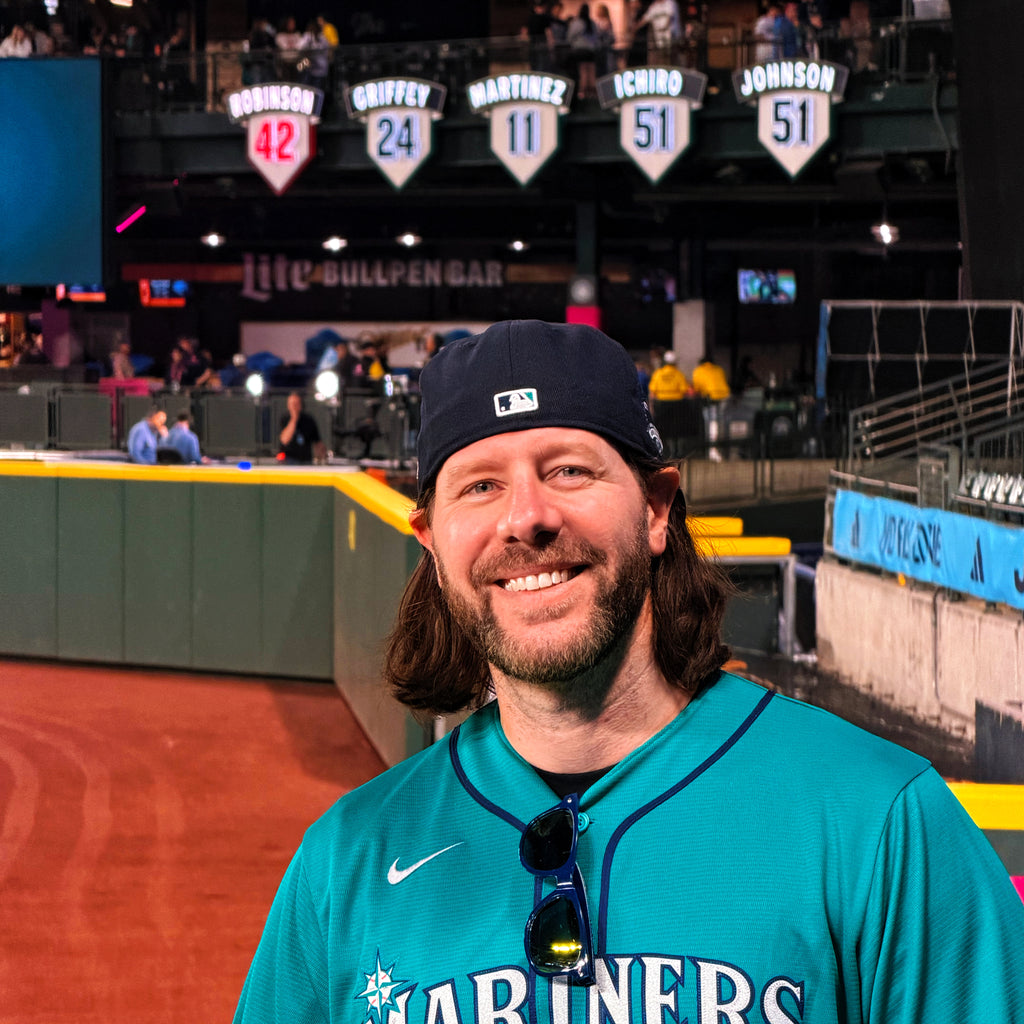Jeremy Deeds wearing a teal Mariners jersey with sunglasses on his shirt, standing in Mariner's baseball stadium.