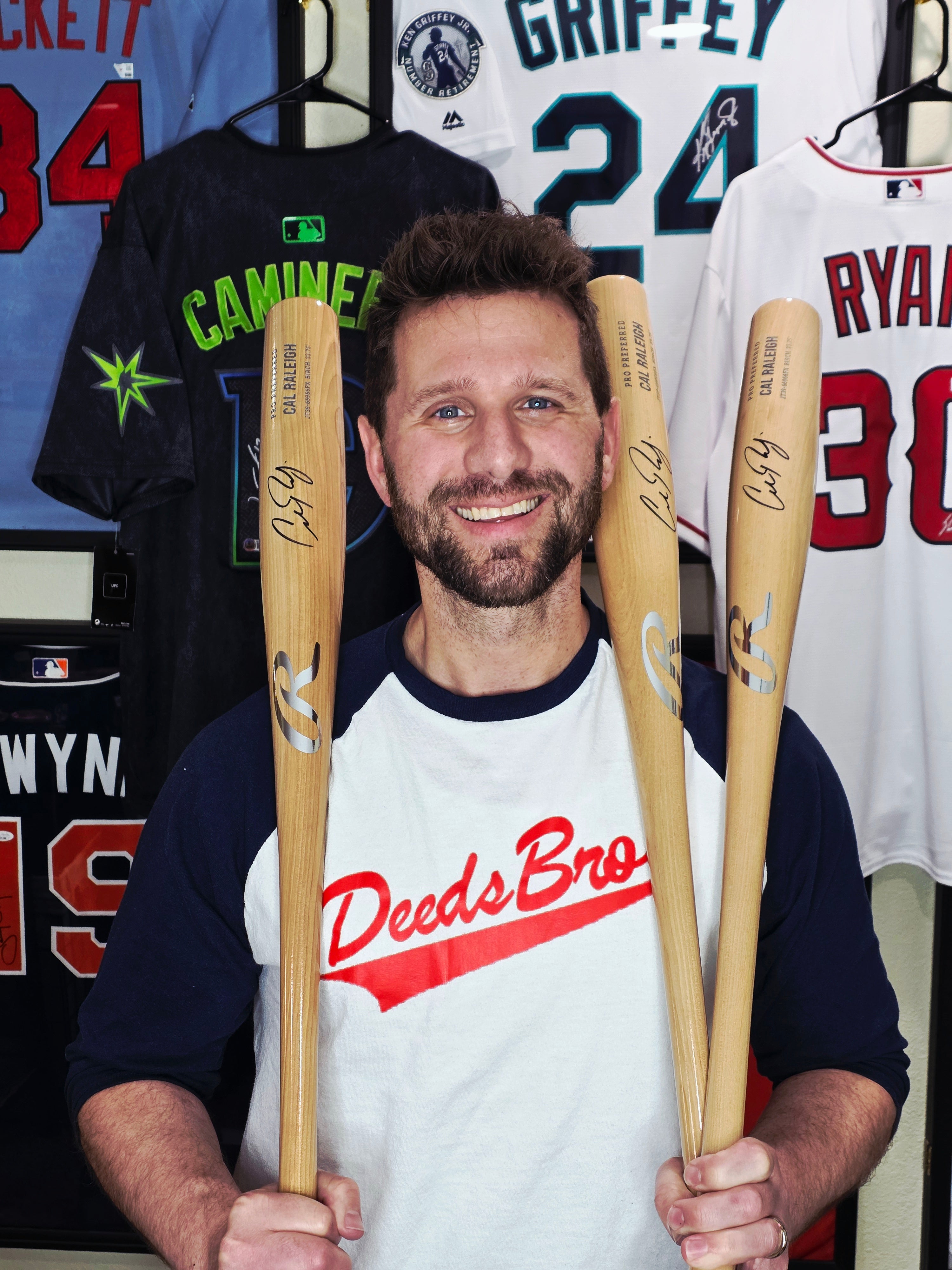 Man holding three baseball bats in front of sports jerseys